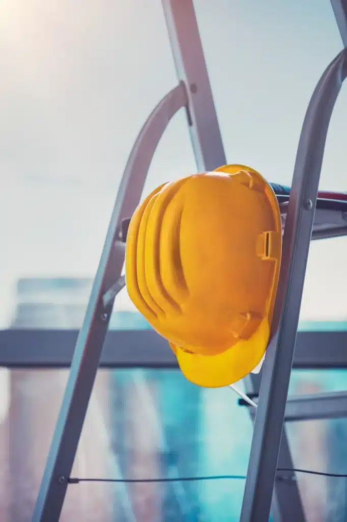 Casque de chantier jaune sur échelle : Sécurité des travaux. Casque de sécurité jaune vif suspendu à un escabeau métallique. Travaux, construction, et skyline urbaine floue en arrière-plan.