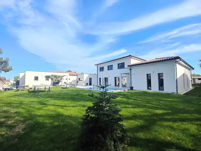 Villa moderne avec piscine et grand jardin sous un ciel bleu Façade arrière d'une maison moderne blanche à deux étages avec toit tuilé, donnant sur une piscine et un vaste jardin sous un ciel bleu.