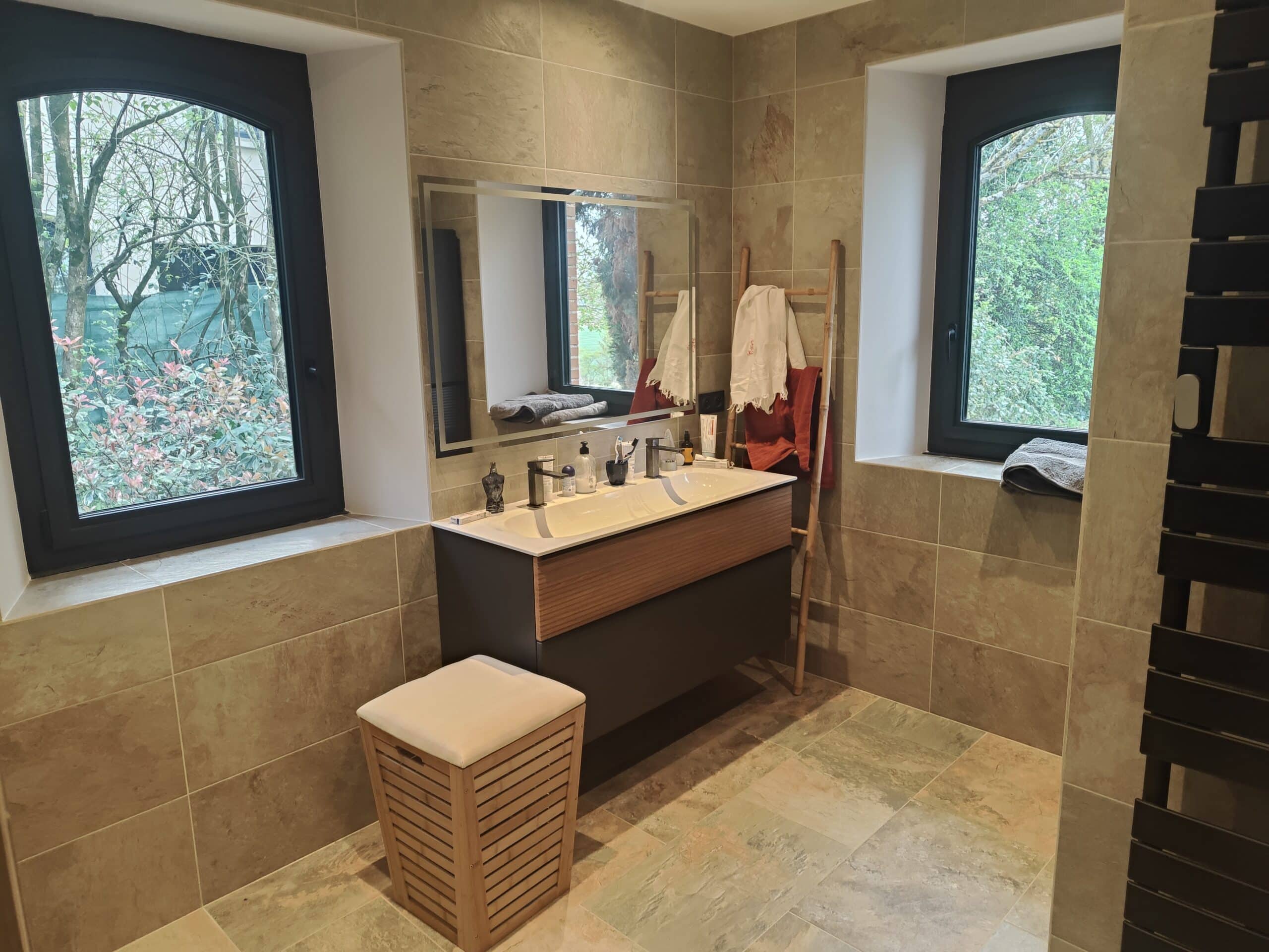 Salle de bain double vasque moderne avec vue nature. Salle de bain moderne : double vasque, grand miroir, carrelage beige texturé. Lumière naturelle par deux fenêtres cintrées sur la verdure.